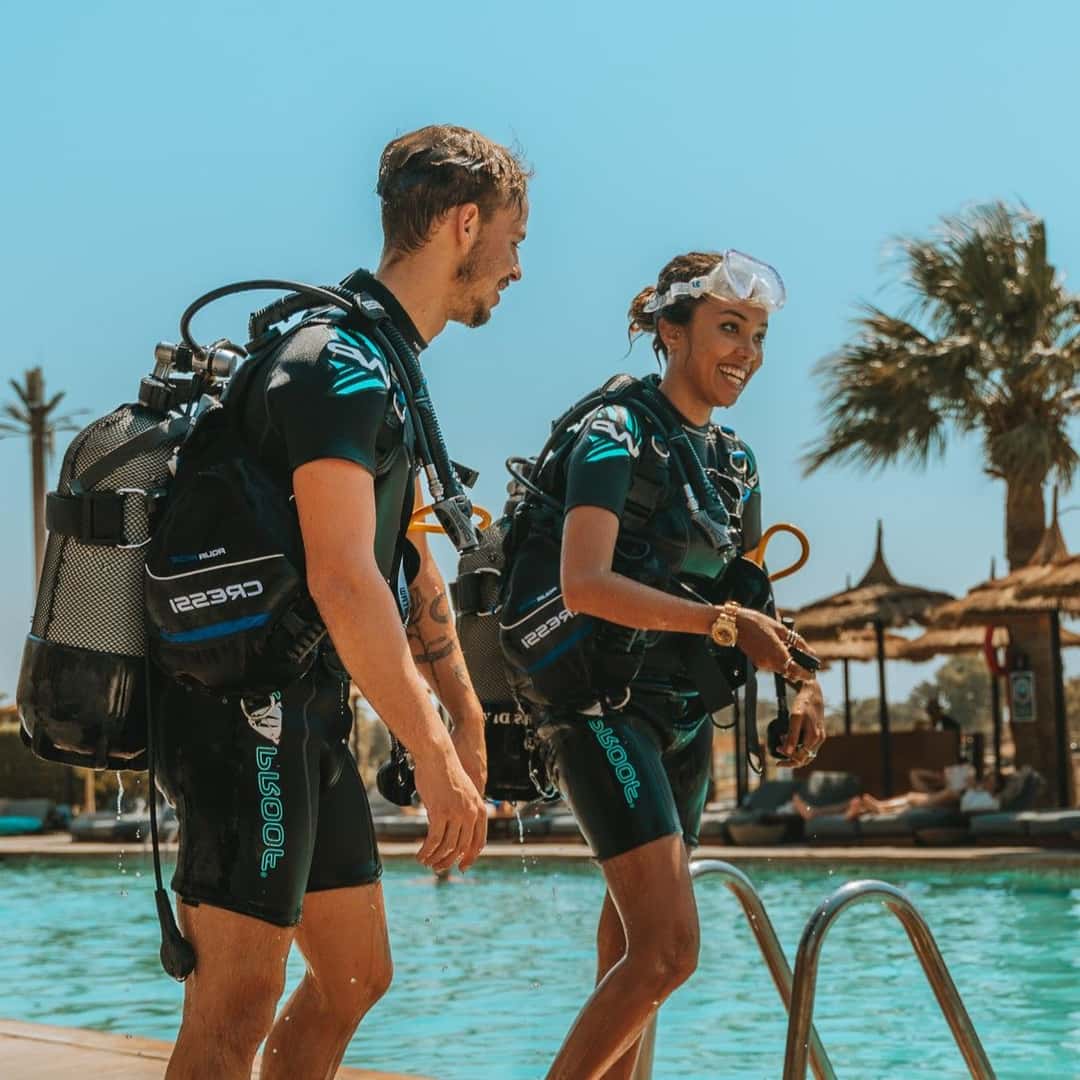 Two divers in full scuba gear stand beside a swimming pool under sunny palm trees.