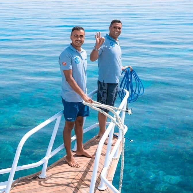 Two smiling men on a boat's bow over clear blue ocean water, one waving.