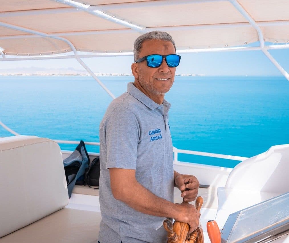 Man in blue sunglasses steering a boat on a bright blue ocean.