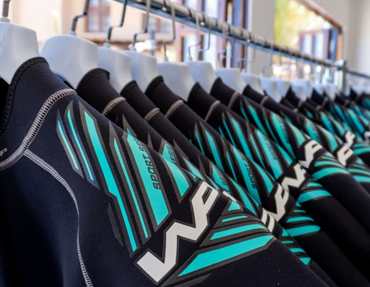 Row of black wetsuits with turquoise and white designs hanging neatly on a retail rack.