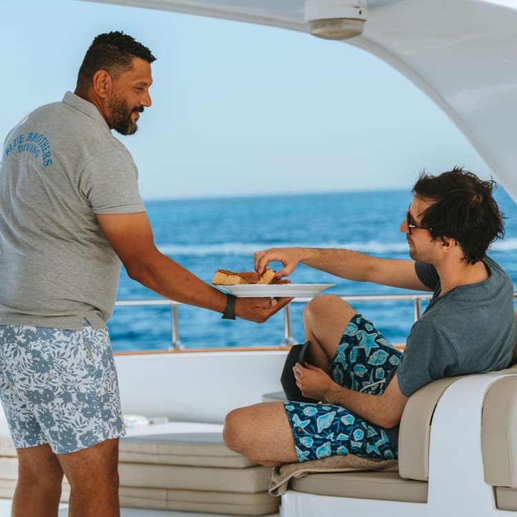 Crew member serves snacks to a guest enjoying a sunny day on a yacht.