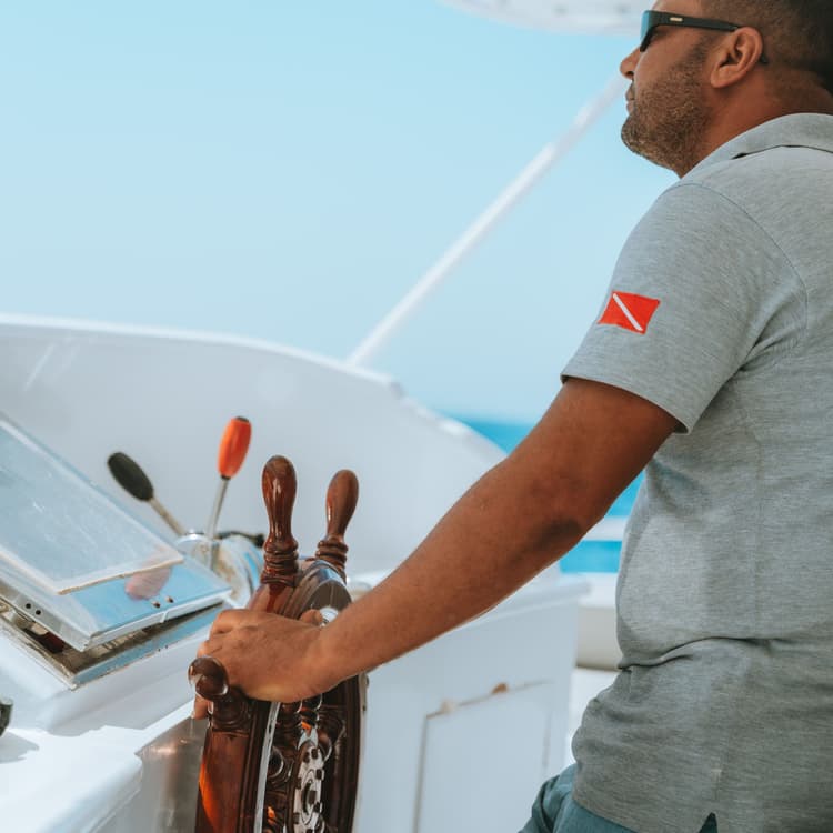 Man in sunglasses steers a boat with a wooden helm under a clear blue sky.
