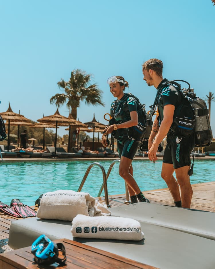 Two divers carrying full scuba gear walking towards a swimming pool for a training session.