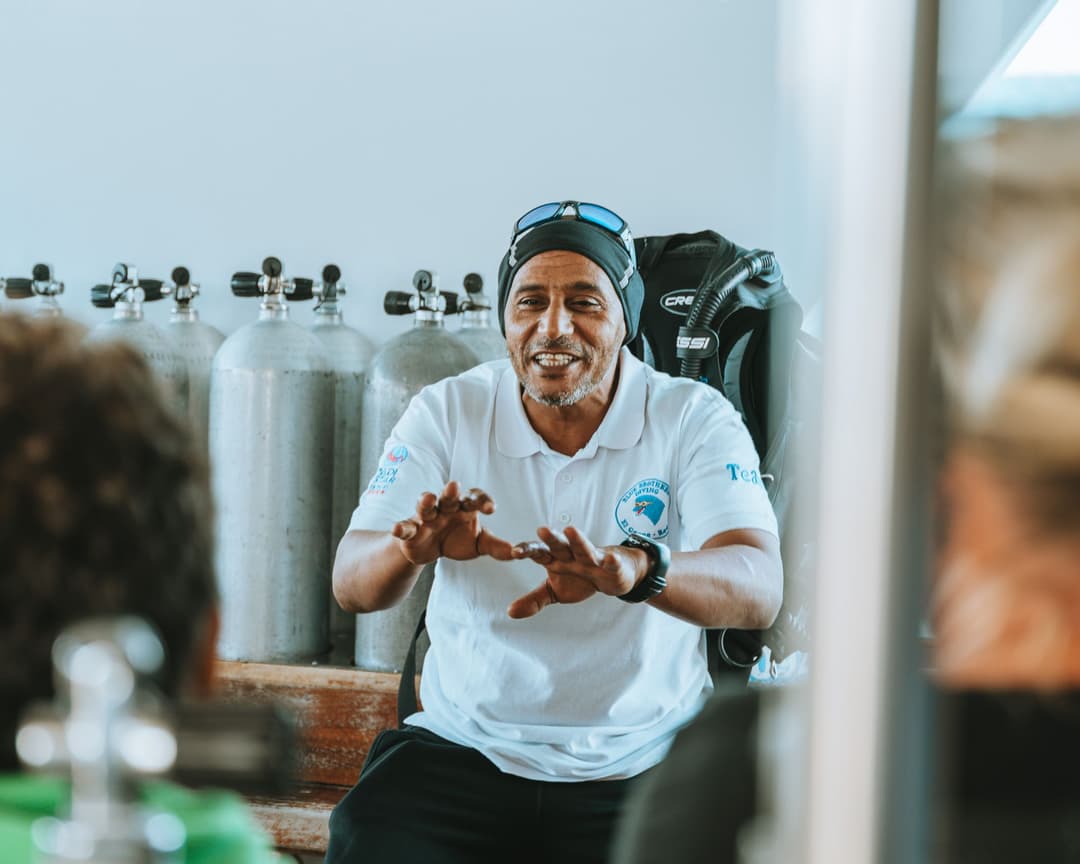 A dive instructor speaking to a group of students in an indoor classroom setting, with scuba tanks behind him.