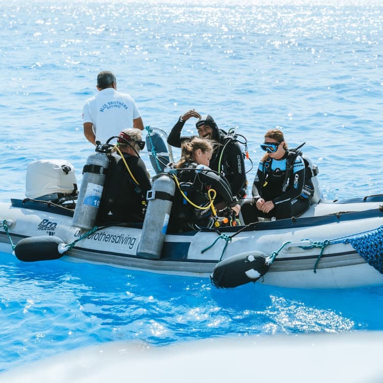 Divers in wetsuits and full gear sit in an inflatable boat on clear blue water.