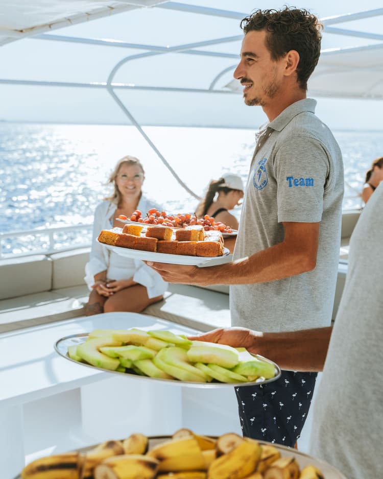 A man smiling and serving trays of food to guests seated on a boat.