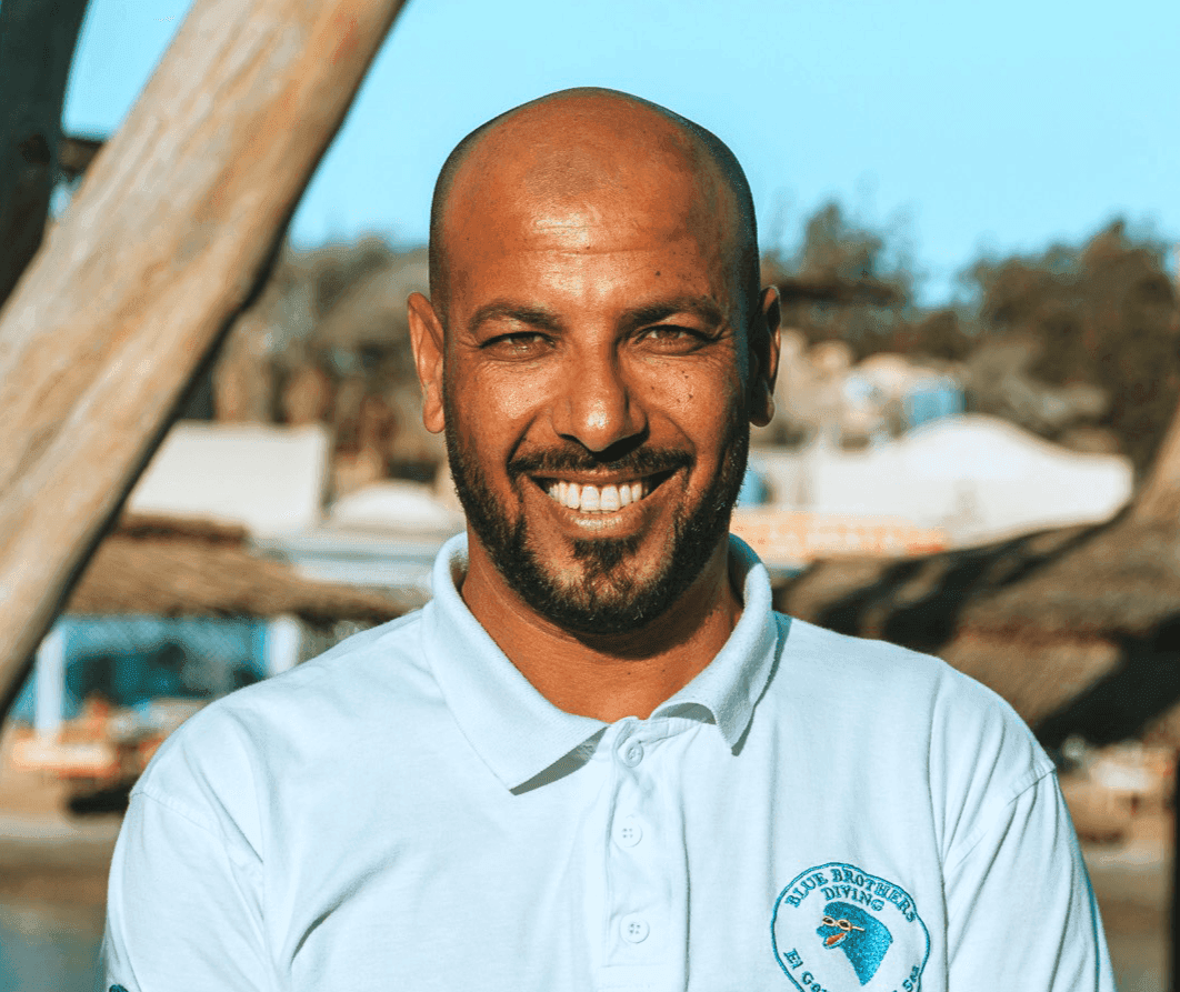 Bald man with a beard smiles, wearing a white polo shirt outdoors.