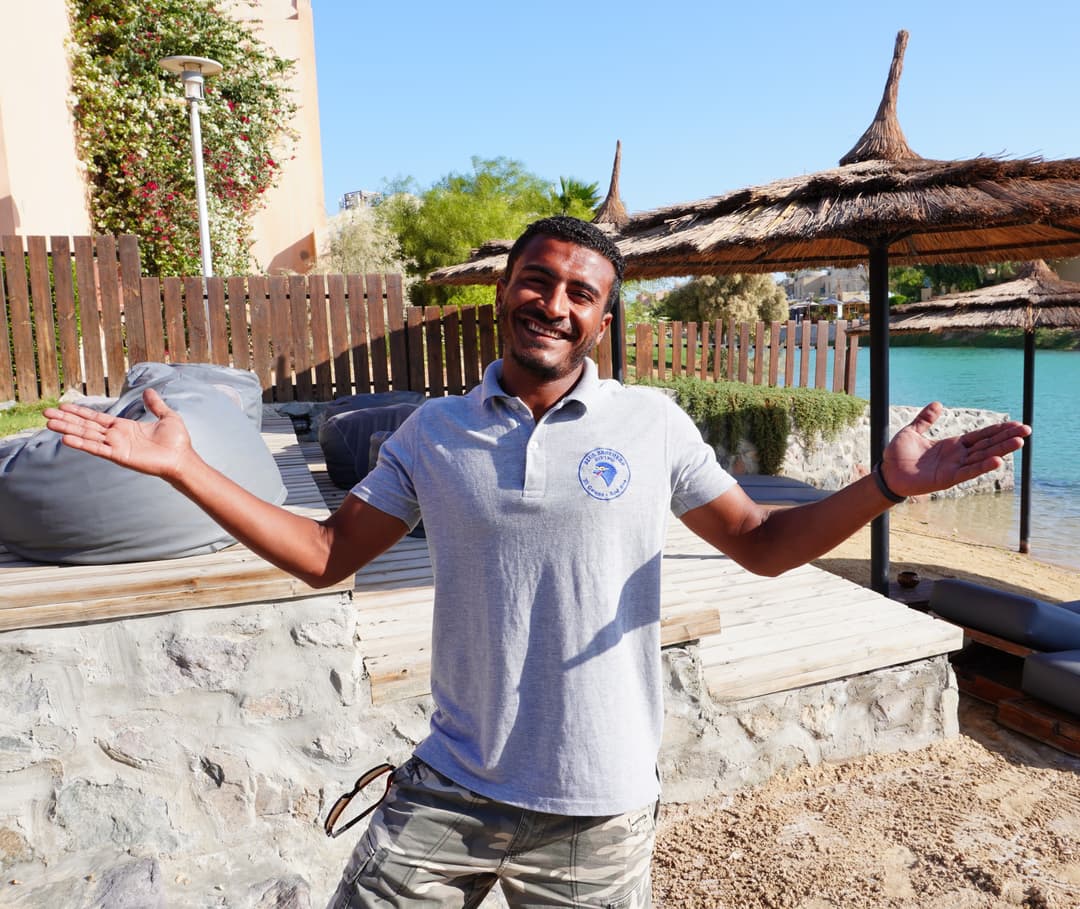 A smiling man with outstretched arms stands outdoors at a sunny resort with water.