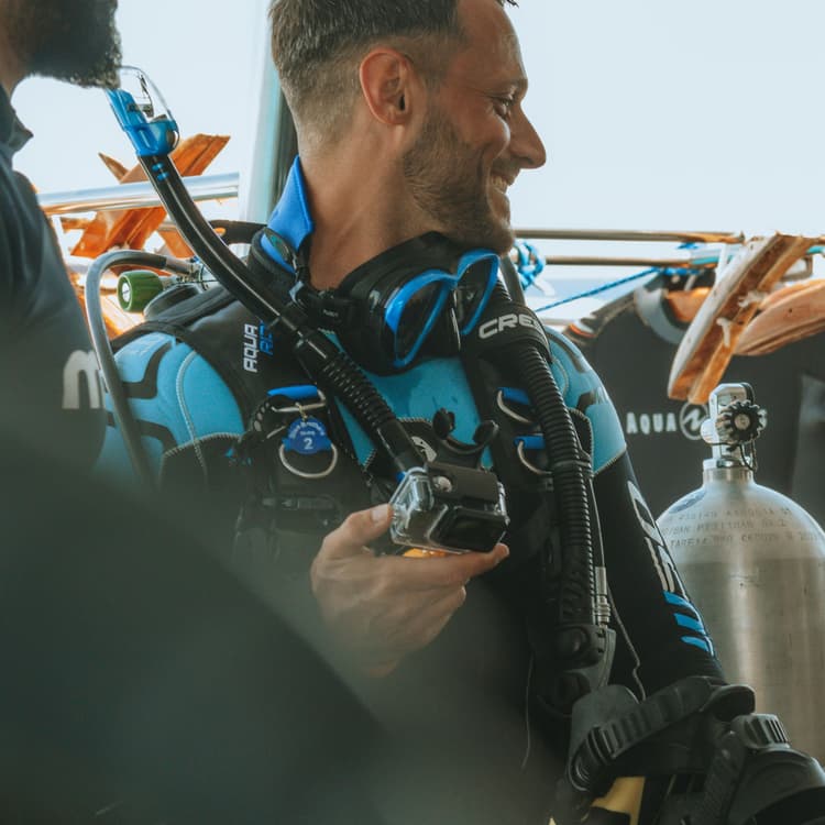 A smiling man in blue and black scuba gear holds diving equipment on a boat.
