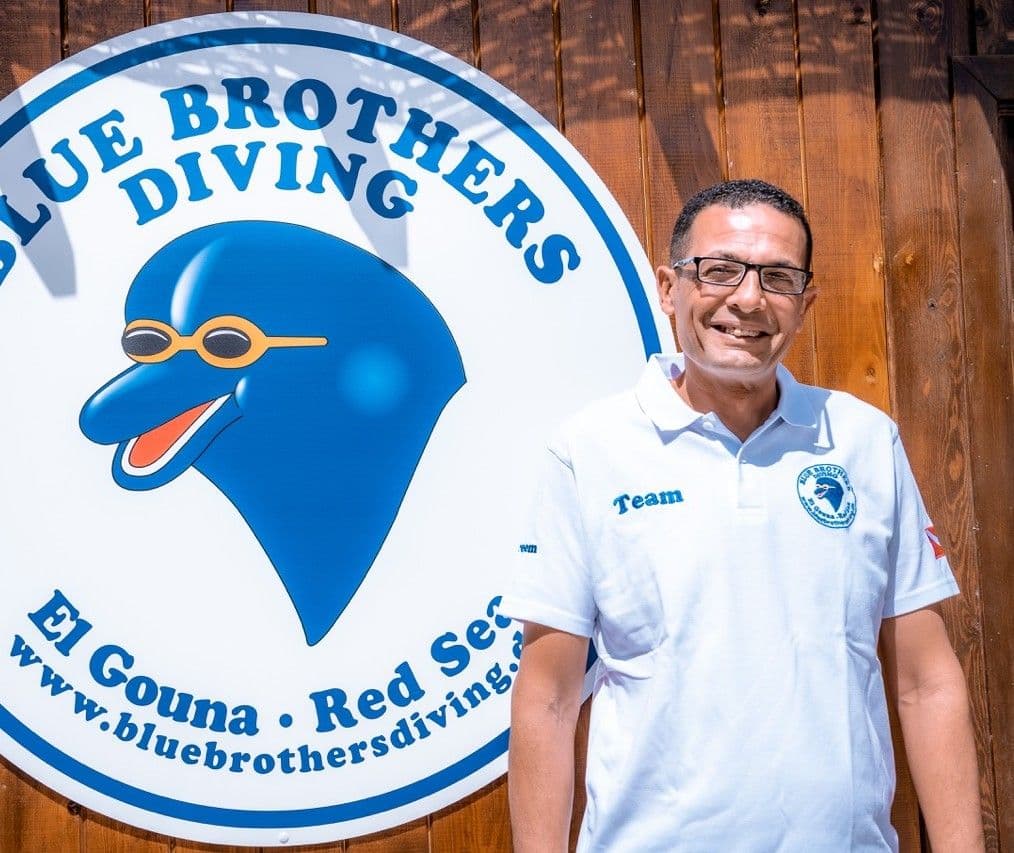 Smiling man in white polo shirt stands next to a Blue Brothers Diving sign.