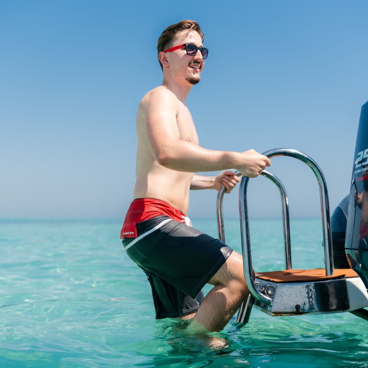 Man in swim trunks and sunglasses climbing a boat ladder from clear blue water.