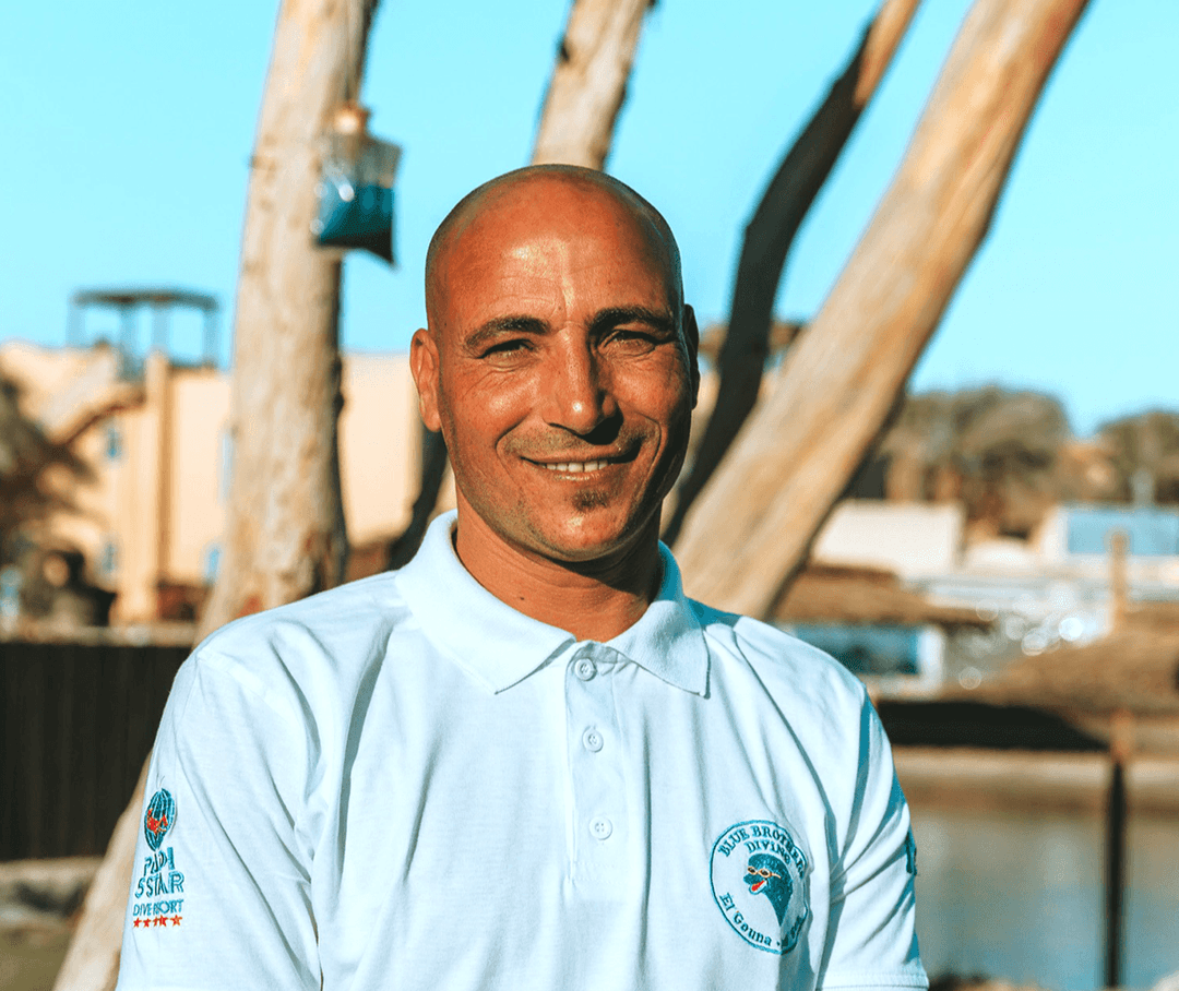 Smiling bald man in a white polo shirt outdoors with trees and buildings.