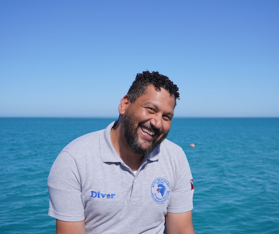 Smiling man in a grey polo shirt with 'Diver' text against a blue ocean background.