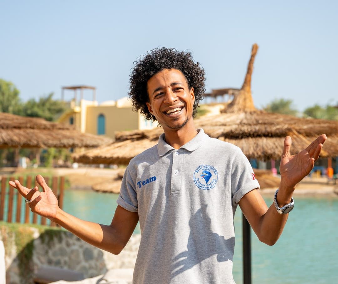 Smiling man in grey polo shirt with outstretched arms in a sunny resort.