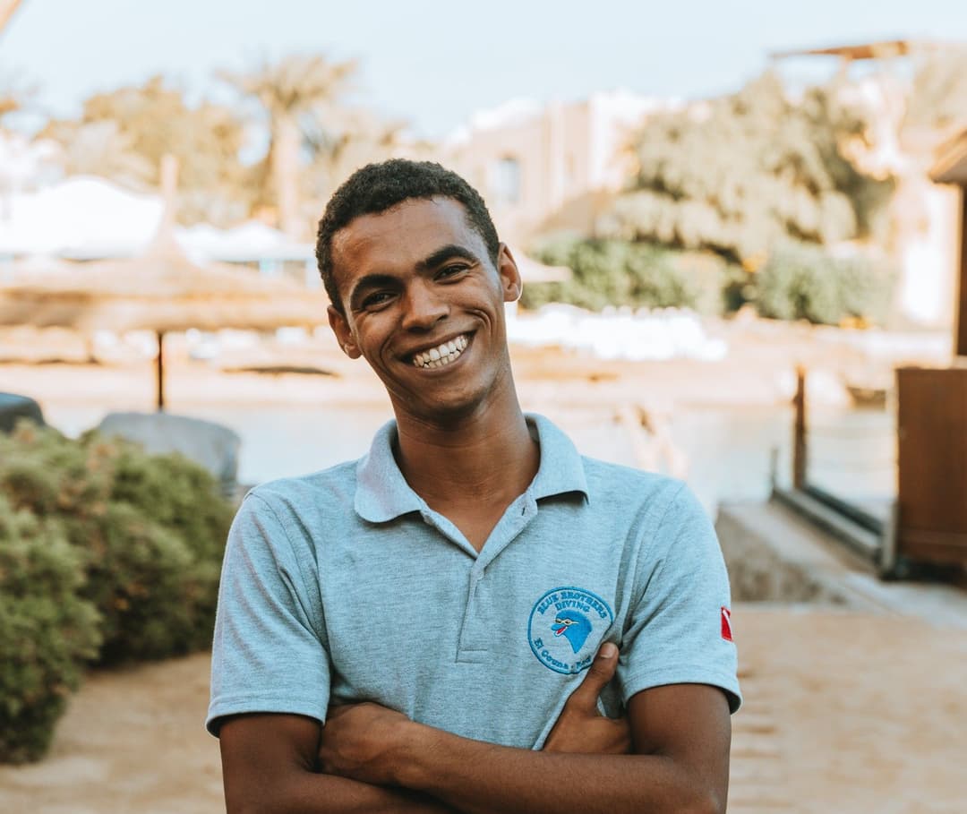 Young man in grey polo shirt smiling with arms crossed at an outdoor resort.